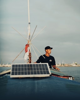 A man stands on a boat, holding onto its structure, with a large solar panel mounted at the front. The sky is overcast with subtle shades of grey. The background shows a distant shoreline with various structures visible.