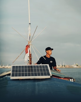 A man stands on a boat, holding onto its structure, with a large solar panel mounted at the front. The sky is overcast with subtle shades of grey. The background shows a distant shoreline with various structures visible.