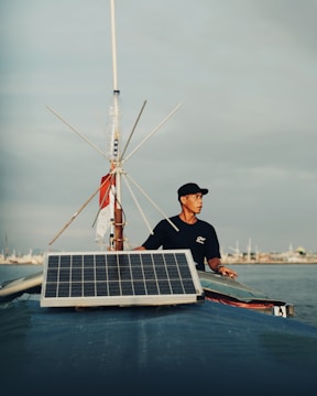 A man stands on a boat, holding onto its structure, with a large solar panel mounted at the front. The sky is overcast with subtle shades of grey. The background shows a distant shoreline with various structures visible.