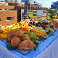 A lavish spread of food arranged on a table, featuring croissants, sliced fruits like oranges and grapes, strawberries, deviled eggs, and various vegetables. The table is adorned with a white tablecloth, and it is set in an outdoor setting with a view of an urban backdrop including buildings and a sports field.