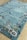A rectangular rug with floral patterns in various colors, primarily set on a light blue background, rests on a light wooden floor. The flowers are stylized and arranged in a border design around the rug's edges, incorporating shades of red, blue, yellow, and brown.