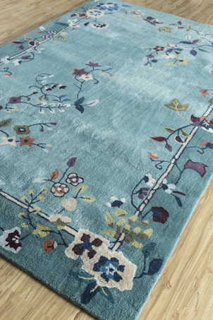 A rectangular rug with floral patterns in various colors, primarily set on a light blue background, rests on a light wooden floor. The flowers are stylized and arranged in a border design around the rug's edges, incorporating shades of red, blue, yellow, and brown.