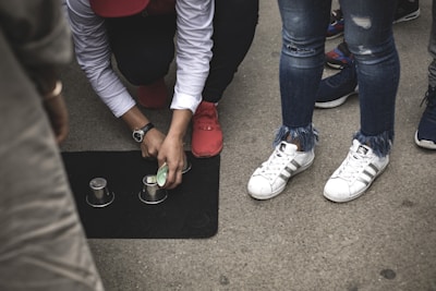 A person crouching on the ground is overseeing a street game involving three metallic cups placed on a dark mat and a small object that resembles a ball. Nearby, several people are watching, and only their legs and shoes are visible.