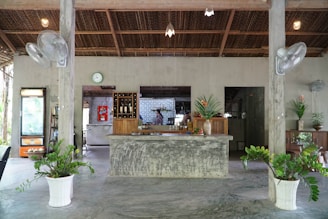 A rustic, open-air cafe interior with a central concrete counter decorated with large vases of tropical plants. The roofing is made of woven materials, and there are ceiling fans and potted plants nearby. In the background, shelves display bottles, and a refrigerator and some furniture are visible.