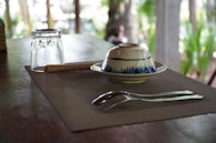 A dining table setup with a placemat featuring a fork and knife, an upside-down glass, a rolled straw or wicker object, and a ceramic bowl with a blue pattern. In the background, there is blurred greenery.