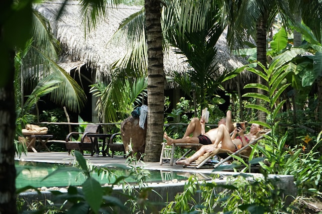Two people are lounging on deck chairs by a lush, tropical poolside area surrounded by dense greenery and palm trees. Their relaxed posture suggests they are enjoying a leisurely day. Behind them, there is a structure with a thatched roof, enhancing the tropical ambiance.