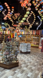 A vibrant store interior filled with colorful, ornate hanging lamps. The ceiling is adorned with intricately designed mosaic lamps in various colors like red, blue, green, and yellow. Below, there are displays of traditional decorative items, ceramics, textiles, and various fabrics stacked neatly. The atmosphere is rich with an array of cultural artifacts and vivid colors.