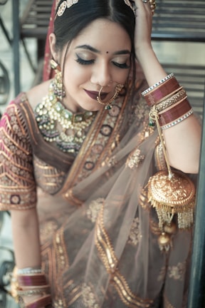 A woman adorned in traditional Indian attire, featuring an ornate saree with intricate gold embroidery. She wears matching jewelry including a large nose ring, bangles, and a statement necklace. Her makeup is elegantly done with heavy eyeliner and dark lipstick. She appears serene and introspective, with one hand resting behind her head.