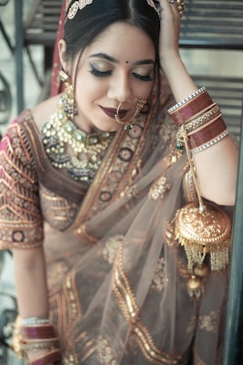 A woman adorned in traditional Indian attire, featuring an ornate saree with intricate gold embroidery. She wears matching jewelry including a large nose ring, bangles, and a statement necklace. Her makeup is elegantly done with heavy eyeliner and dark lipstick. She appears serene and introspective, with one hand resting behind her head.