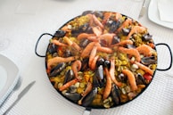 A large pan filled with vibrant paella, featuring cooked prawns, mussels, clams, and vegetables mixed with saffron-infused rice. The seafood is arranged on top, creating an appealing and appetizing look. The dish is placed on a white textured tablecloth, with plates and cutlery neatly arranged around it.