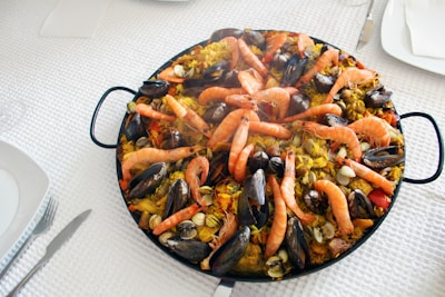 A large pan filled with vibrant paella, featuring cooked prawns, mussels, clams, and vegetables mixed with saffron-infused rice. The seafood is arranged on top, creating an appealing and appetizing look. The dish is placed on a white textured tablecloth, with plates and cutlery neatly arranged around it.