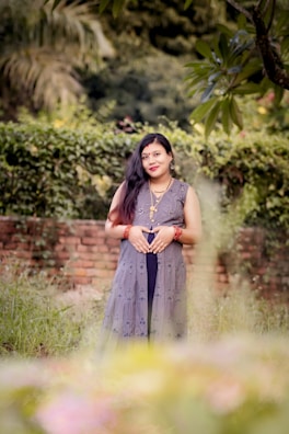 A woman stands in a garden setting with lush greenery and a brick wall in the background. She is wearing a traditional dress with intricate patterns and jewelry, including earrings and bracelets. Her hands are gently placed on her abdomen, and she has long dark hair.