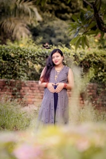 A woman stands in a garden setting with lush greenery and a brick wall in the background. She is wearing a traditional dress with intricate patterns and jewelry, including earrings and bracelets. Her hands are gently placed on her abdomen, and she has long dark hair.