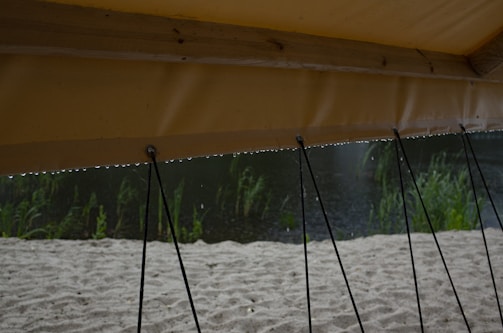 A section of an outdoor canopy is visible, held up by several ropes attached to the ground. Raindrops can be seen on the edge of the canopy above the sand. In the background, tall grass and a body of water are partially visible.