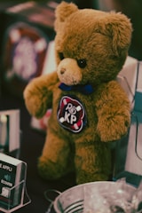 A brown teddy bear with a black nose and dark eyes is seen standing upright. The bear has a blue bow tie around its neck and a circular patch on its chest with letters 'BS KP' and small white objects resembling pills on the patch. The background is blurred, but there appear to be other objects or products around the bear.