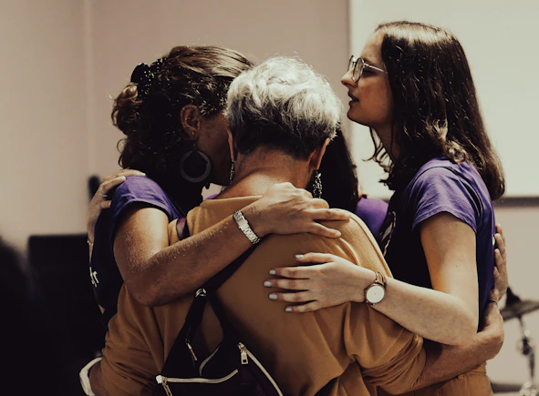 A group of people standing close together in an embrace, showing a sense of unity and support. The individuals are wearing casual clothing and the setting appears to be indoors, possibly in a social or community event.