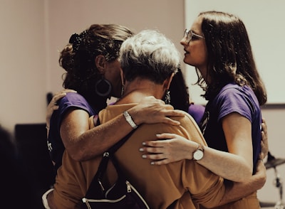 A group of people standing close together in an embrace, showing a sense of unity and support. The individuals are wearing casual clothing and the setting appears to be indoors, possibly in a social or community event.