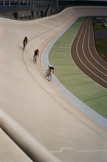 Three cyclists are riding on an outdoor velodrome, navigating a steeply banked curve. The track is marked with lines of blue, red, and other colors, and the setting appears to be an overcast day.