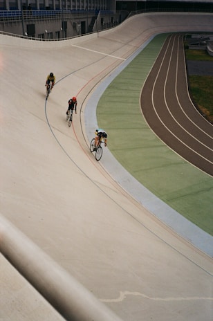 Three cyclists are riding on an outdoor velodrome, navigating a steeply banked curve. The track is marked with lines of blue, red, and other colors, and the setting appears to be an overcast day.