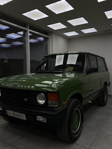 A vintage Range Rover in a rich green color is displayed in a showroom with modern lighting. The vehicle is parked on a tiled floor next to large windows reflecting the ceiling lights.