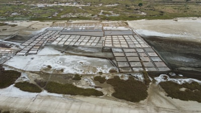 Aerial view of a large expanse of square salt pans arranged in a grid pattern. The area surrounding the pans is patchy with salt deposits and sparse vegetation, lying adjacent to uneven terrain. Lush greenery is visible in the distant background.