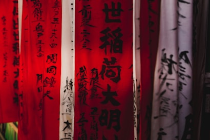Vertical banners with Japanese calligraphy hang closely together, featuring red and white backgrounds with black characters. The inscriptions appear to be traditional and are artistically styled.