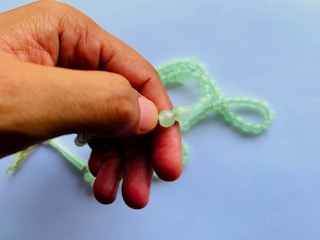A hand is holding a piece of green prayer beads or a rosary against a soft blue background. The beads are translucent, possibly made of glass or jade, and they are loosely coiled in the background.