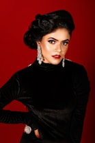 A woman with vintage-style hair and makeup poses against a deep red background. She wears a black velvet dress and large, ornate silver earrings. Her makeup is dramatic, with bold red lipstick and expressive eye makeup. Her pose is confident, with one hand on her hip and the other resting by her side.