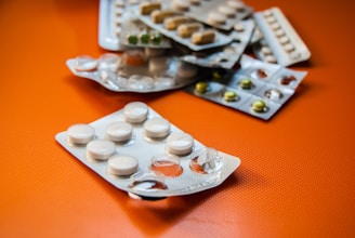 A collection of various blister packs containing different types of tablets and capsules are spread out on an orange surface. The blister packs are partially empty, with some tablets missing.