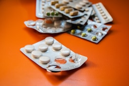 A collection of various blister packs containing different types of tablets and capsules are spread out on an orange surface. The blister packs are partially empty, with some tablets missing.