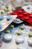Various blister packs containing pills are displayed, featuring different colors such as white, green, blue, and red. The image focuses on the texture and layout of the blister packs scattered around.