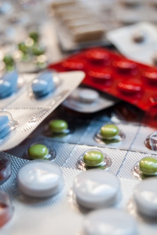 Various blister packs containing pills are displayed, featuring different colors such as white, green, blue, and red. The image focuses on the texture and layout of the blister packs scattered around.