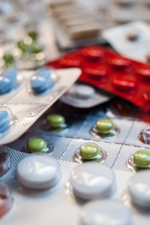 Various blister packs containing pills are displayed, featuring different colors such as white, green, blue, and red. The image focuses on the texture and layout of the blister packs scattered around.