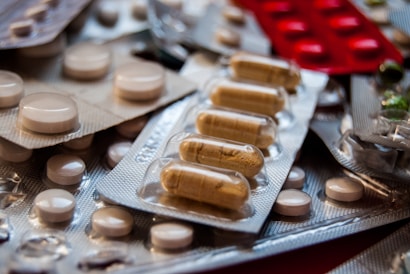 A close-up view of various pills and capsules in blister packs, including brown capsules and white round tablets. The blister packs are arranged haphazardly, with some lying flat and others overlapping. The lighting highlights the shiny surface of the packaging, creating reflections.