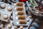 A variety of medications are arranged in blister packs, featuring different shapes and colors, including round white tablets and oblong brown capsules. The packs are overlapping, creating a textured surface.