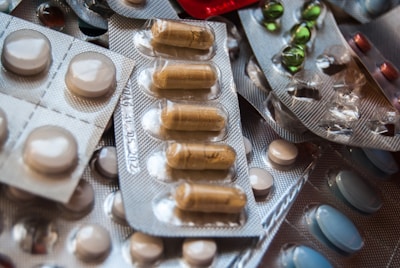 A variety of medications are arranged in blister packs, featuring different shapes and colors, including round white tablets and oblong brown capsules. The packs are overlapping, creating a textured surface.