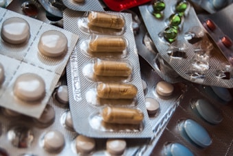 A variety of medications are arranged in blister packs, featuring different shapes and colors, including round white tablets and oblong brown capsules. The packs are overlapping, creating a textured surface.