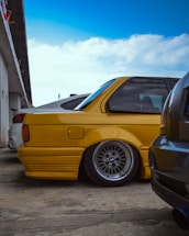 A bright yellow car with a lowered suspension is prominently displayed. It features stylish, intricate alloy wheels and is parked on a concrete surface. The background includes a partial view of a building with people observing the vehicles.