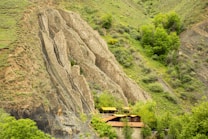 A rustic wooden house nestled at the base of a large, rugged hillside covered with sparse grass and lush green trees. The hillside features distinctive geological rock formations that jut out dramatically. The scene blends natural elements with a simple human habitation, creating a serene landscape.