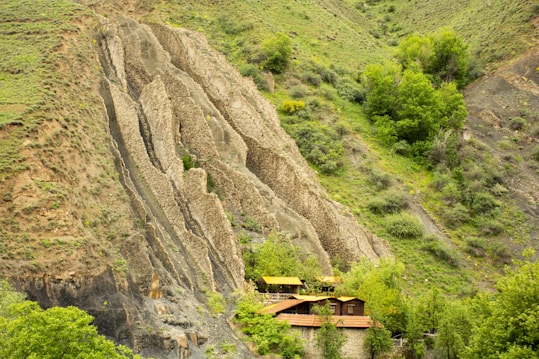 A rustic wooden house nestled at the base of a large, rugged hillside covered with sparse grass and lush green trees. The hillside features distinctive geological rock formations that jut out dramatically. The scene blends natural elements with a simple human habitation, creating a serene landscape.