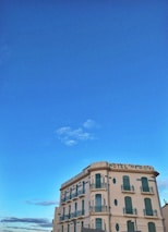 A vintage-style hotel building with several stories, adorned with green shutters and balconies. The structure is situated under a clear blue sky, giving it a picturesque and serene appearance.