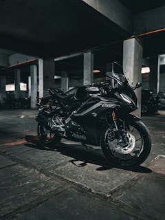 A sleek black Yamaha motorcycle is parked in a dimly lit concrete parking garage. The setting is industrial, with numerous columns and a stone-paved floor visible. The lighting creates a moody, shadow-filled atmosphere, enhancing the motorcycle's reflective surfaces and sharp contours.