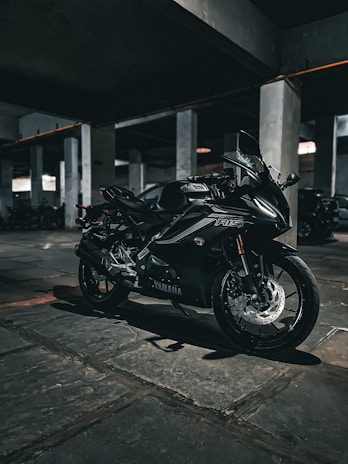 A sleek black Yamaha motorcycle is parked in a dimly lit concrete parking garage. The setting is industrial, with numerous columns and a stone-paved floor visible. The lighting creates a moody, shadow-filled atmosphere, enhancing the motorcycle's reflective surfaces and sharp contours.