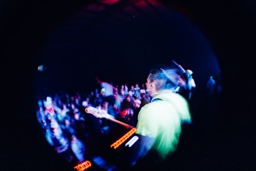 A vibrant concert scene captured with a fisheye lens, showing a musician playing a guitar on stage, surrounded by a lively audience under dynamic lighting.