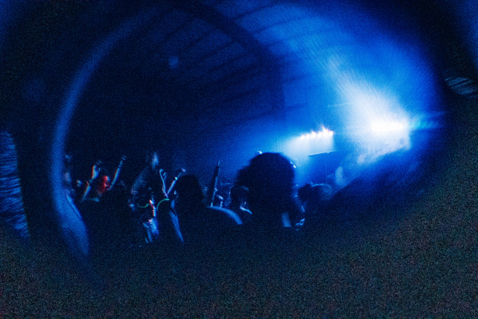 A panoramic shot of a packed venue with light blue and black lighting, showing young people enjoying music and good vibes.
