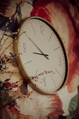 A large round wall clock with a golden rim is mounted against a floral patterned wall. The background features a variety of reds, pinks, and greens, depicting large flower petals. Some small branches with leaves and red buds are visible in the foreground.