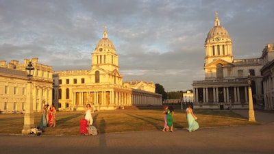 An elegant architectural scene featuring two historic domed buildings with columns, basking in the warm glow of the setting sun. In the foreground, a group of people in colorful dresses stand or walk across an open grassy area, creating a lively contrast against the serene backdrop.