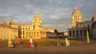 An elegant architectural scene featuring two historic domed buildings with columns, basking in the warm glow of the setting sun. In the foreground, a group of people in colorful dresses stand or walk across an open grassy area, creating a lively contrast against the serene backdrop.