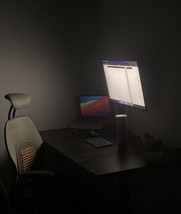 A dimly lit workspace with a dark wooden desk, featuring a modern ergonomic chair with a mesh backrest and lumbar support cushion. A large monitor displays a light-colored application interface, and a laptop with a colorful desktop background is positioned slightly open. On the desk are a keyboard, a mouse, a cylindrical container, and a small plant in a pot.