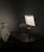 A dimly lit workspace with a dark wooden desk, featuring a modern ergonomic chair with a mesh backrest and lumbar support cushion. A large monitor displays a light-colored application interface, and a laptop with a colorful desktop background is positioned slightly open. On the desk are a keyboard, a mouse, a cylindrical container, and a small plant in a pot.